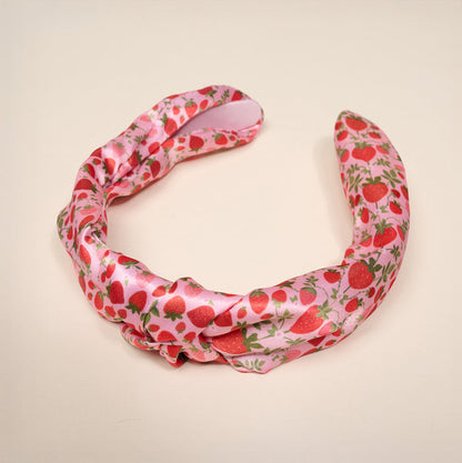 The Darling Effect's Strawberry Fields Classic Twist Headband features pink satin with red strawberries, green leaves, and a charming knot detail, shown on a plain light background.