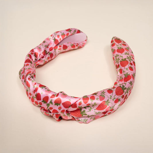 The Darling Effect's Strawberry Fields Classic Twist Headband features pink satin with red strawberries, green leaves, and a charming knot detail, shown on a plain light background.