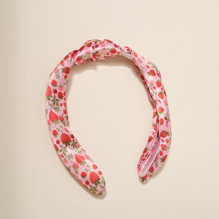 The Darling Effect’s Strawberry Fields Classic Twist Headband features red strawberry and green leaf prints with a stylish knot detail, shown against a beige background—a versatile accessory for any outfit.