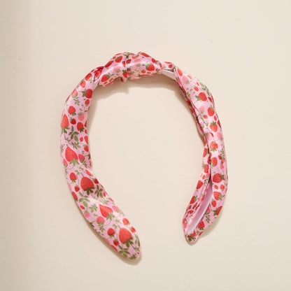 The Darling Effect’s Strawberry Fields Classic Twist Headband features red strawberry and green leaf prints with a stylish knot detail, shown against a beige background—a versatile accessory for any outfit.