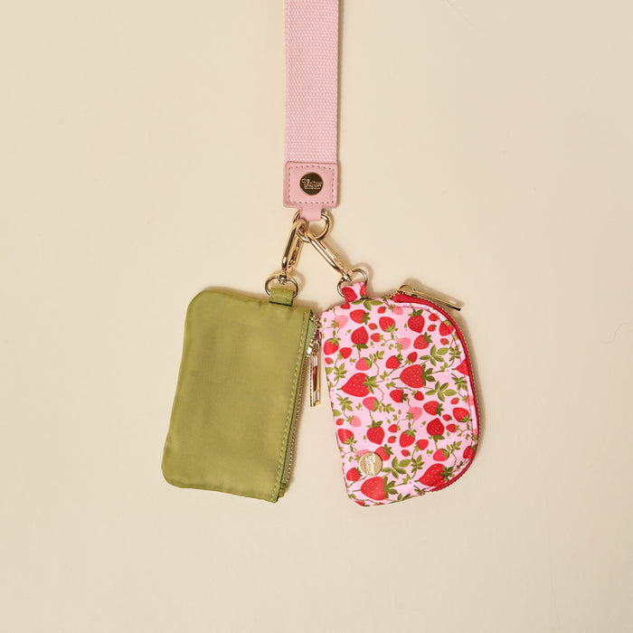 The Darling Effect’s Strawberry Fields Dynamic Duo Pouch Wristlet features a luxe light pink wristlet lanyard with two pouches—one olive green, one white with red strawberries—both attached by gold key rings for stylish, hands-free carrying.
