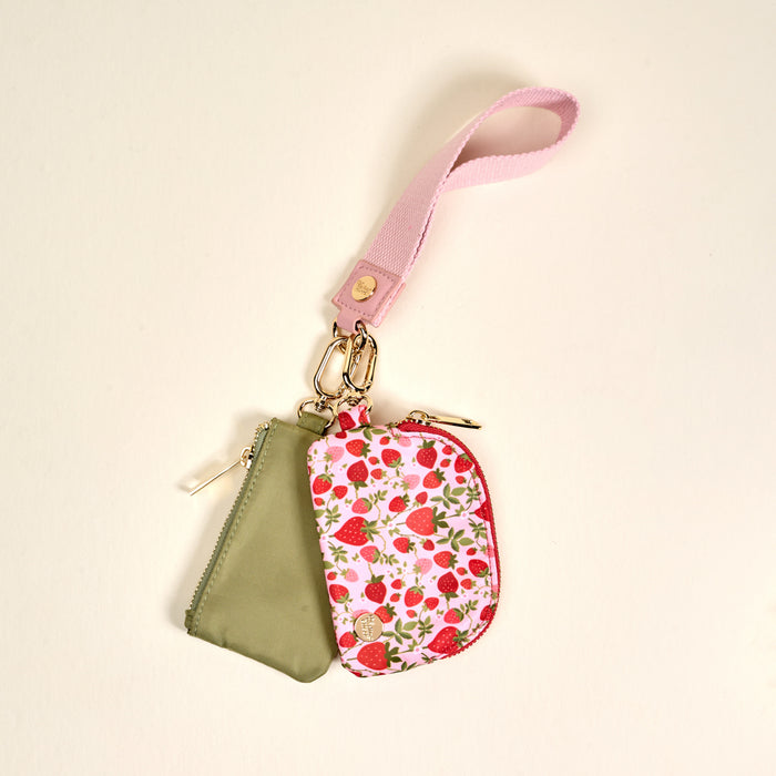 The Darling Effect’s Strawberry Fields Dynamic Duo Pouch Wristlet features two small zippered pouches—one olive green, one white with pink strawberry print—attached by gold hardware to a pink fabric keychain.