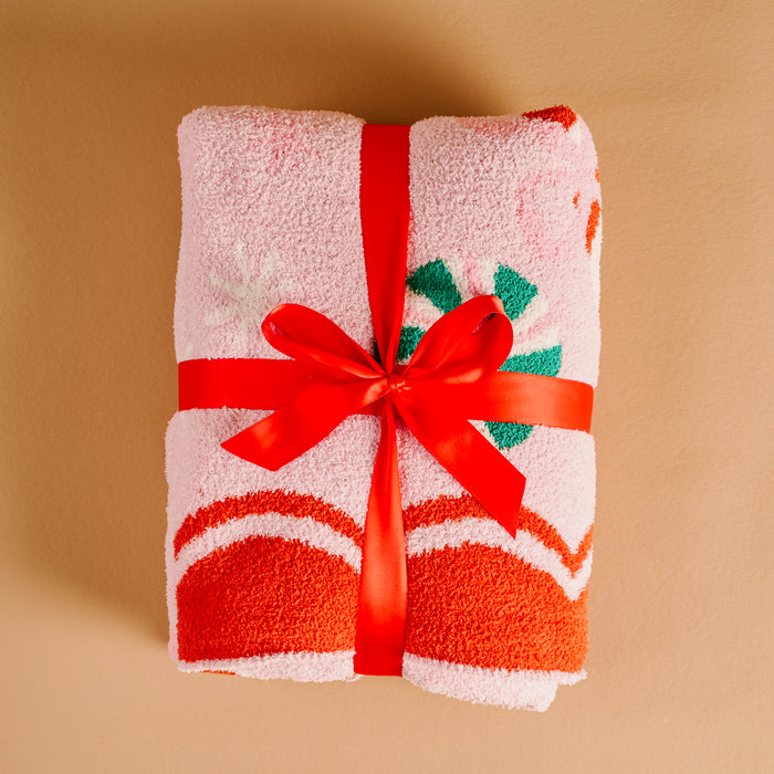 A rolled-up Sugar Rush Cozy Dreams Luxe Blanket by The Darling Effect, featuring a fuzzy pink Christmas pattern and tied with a red ribbon and bow, sits on a tan background.