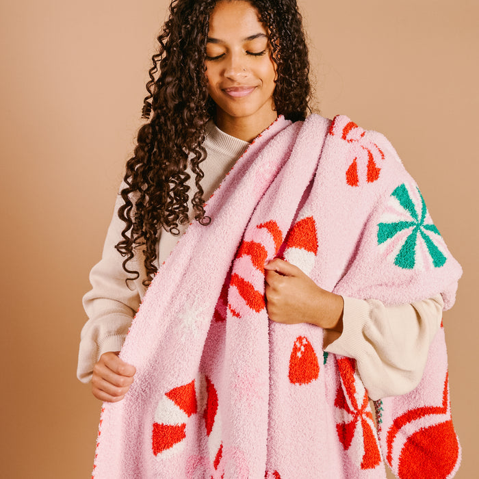 A woman with curly hair, in a beige sweater, smiles gently while wrapped in The Darling Effect Sugar Rush Cozy Dreams Luxe Blanket, featuring red and white candy canes and green peppermint designs, against a plain tan background.