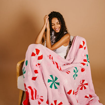 A smiling woman with curly hair sits on a chair, wrapped in The Darling Effect Sugar Rush Cozy Dreams Luxe Blanket—a pink ultra-soft throw with red, green, and white peppermint candy designs—against a plain beige background.