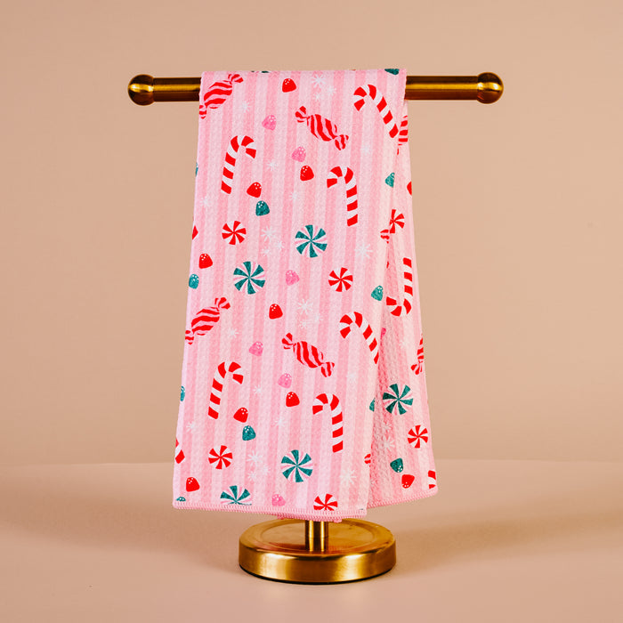 The Darling Effect’s Sugar Rush Waffle Tea Towel, featuring red and white candy canes, red and green peppermint candies, and red hearts on pink, hangs on a gold towel stand against a beige background.