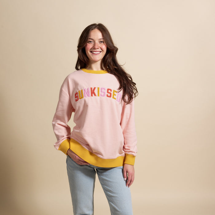 A smiling woman with long brown hair wears The Darling Effect's Sunkissed Oversized Crew in light pink with yellow cuffs, collar, and embroidered details, paired with light blue jeans, standing against a beige background.