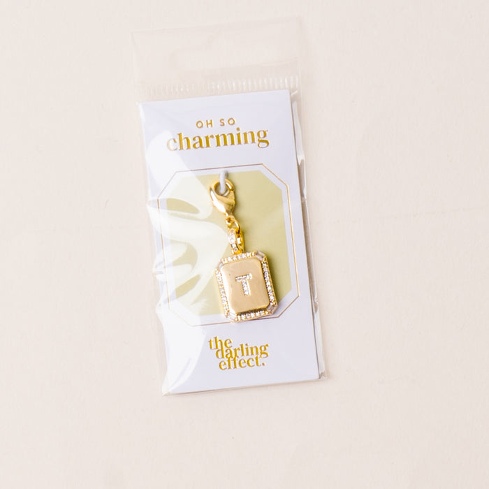 The T Jewelry Charm Initial Letter Gold by the-darling-effect features a gold-plated brass charm with a beveled frame, diamond accents, and a central white T. Packaged with OH SO charming and the darling effect logos.