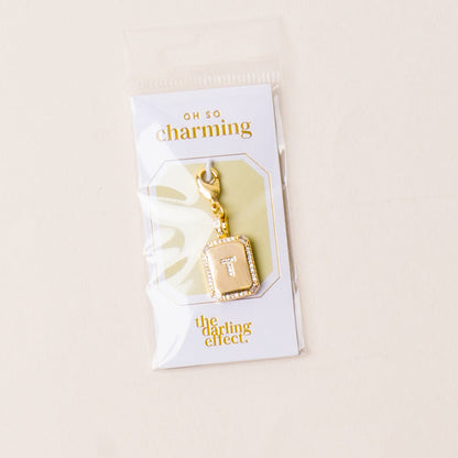 The T Jewelry Charm Initial Letter Gold by the-darling-effect features a gold-plated brass charm with a beveled frame, diamond accents, and a central white T. Packaged with OH SO charming and the darling effect logos.