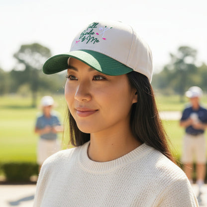 A young woman smiles outdoors on a sunny day, wearing The Darling Effect's Talk Birdie to Me Classic Canvas Hat in green and white, with people and a golf course visible in the blurred background.