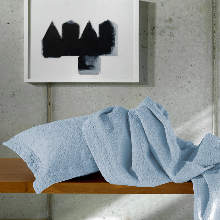 Tamara-Blue Decorative pillowcase and bedspread draped over a wooden surface with abstract art in the background.