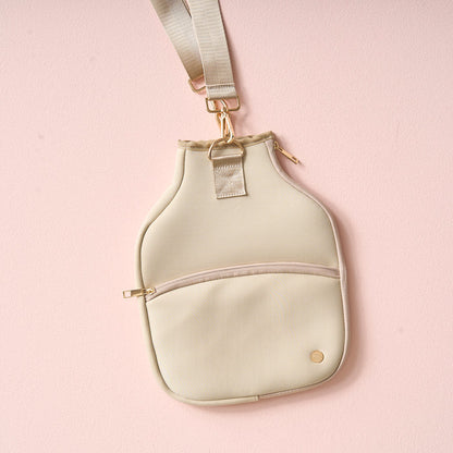 The Darling Effect Tan Pickleball Paddle Case, shown against a light pink background, features gold zippers and hardware, an adjustable strap, a front zip pocket, and a round gold logo—ideal for carrying your pickleball essentials.