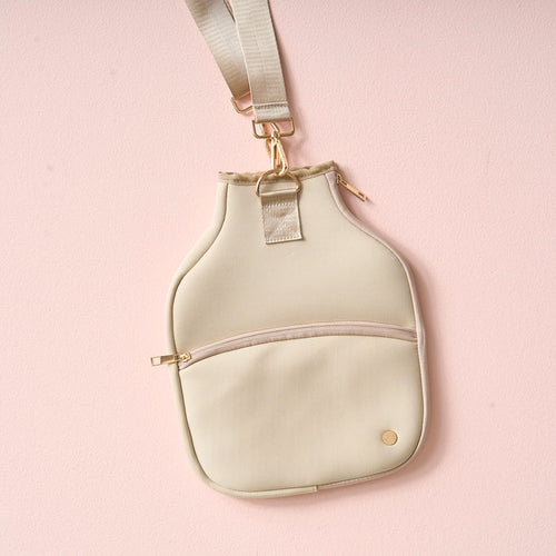 The Darling Effect Tan Pickleball Paddle Case, shown against a light pink background, features gold zippers and hardware, an adjustable strap, a front zip pocket, and a round gold logo—ideal for carrying your pickleball essentials.