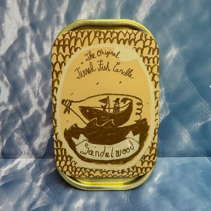The Original Tinned Fish Candle in Sandalwood scent by Tinned Candle on blue patterned surface with cute fish illustration.
