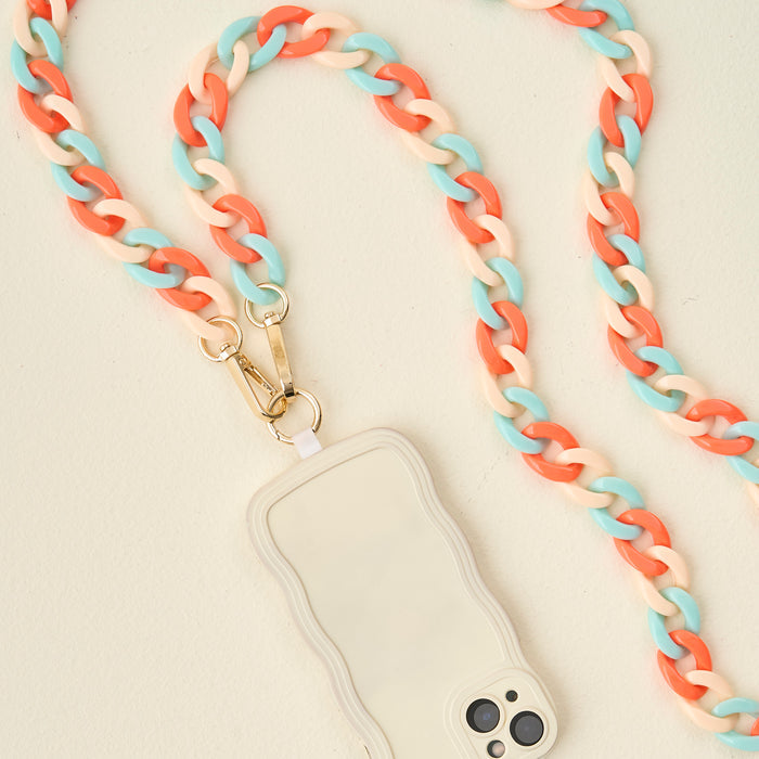 The Darling Effect’s Tri Coastal Hold the Phone Crossbody Chain pairs a multicolored coral, blue, and beige chain with a wavy-edged cream case, making this standout accessory pop against a light cream background.