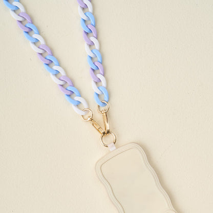The Darling Effect's Tri Cool Hold the Phone Crossbody Chain features pastel blue, purple, and white links with gold clasps—an on-trend accessory for your cream, wavy-edged badge holder or phone, styled on a light beige surface.