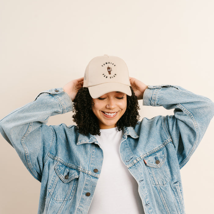 A smiling woman with curly hair wears The Darling Effect's Tumbler Fan Club Trckr Hat, which features stylish text and a tumbler graphic, paired with a denim jacket.