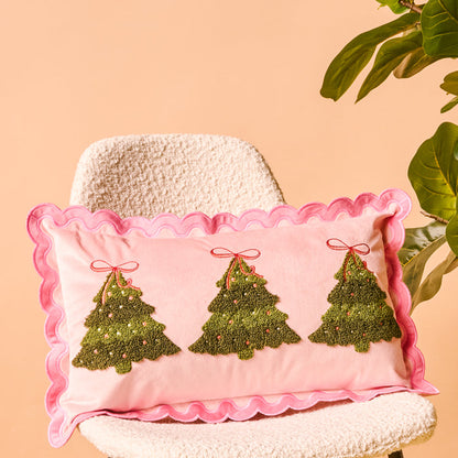The Darling Effect's Twinkling Trees Scalloped Rectangle Throw Pillow, in pink with ruffled edges and embroidered trees, brings cozy holiday charm to decor when placed on a textured chair near greenery against a peach background.