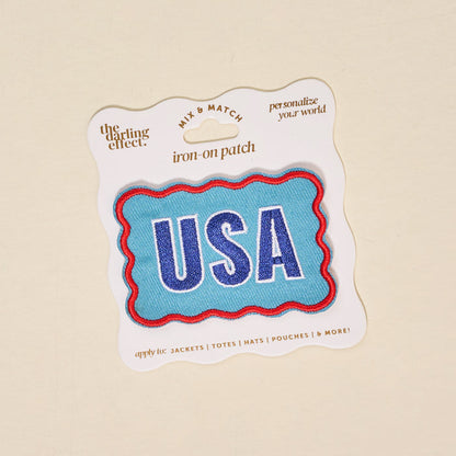 The Darling Effect's USA Mix & Match Iron-On Patch features bold white and blue USA letters with a red wavy border, packaged for easy gifting—ideal for customizing jackets with patriotic flair.