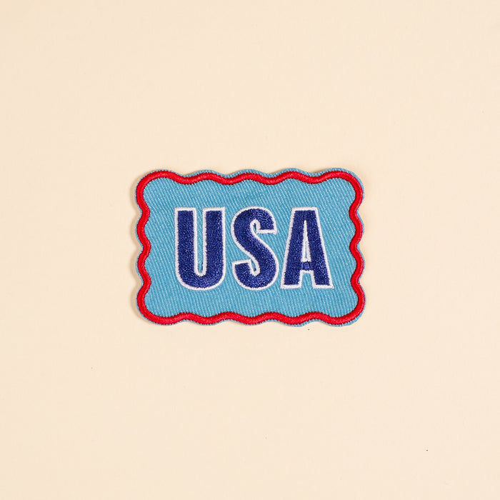The Darling Effect’s USA Mix & Match Iron-On Patch features bold blue and white “USA” letters with a red wavy border on beige—ideal for customizing jackets or adding to your colorful patch collection.