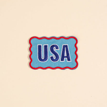 The Darling Effect’s USA Mix & Match Iron-On Patch features bold blue and white “USA” letters with a red wavy border on beige—ideal for customizing jackets or adding to your colorful patch collection.