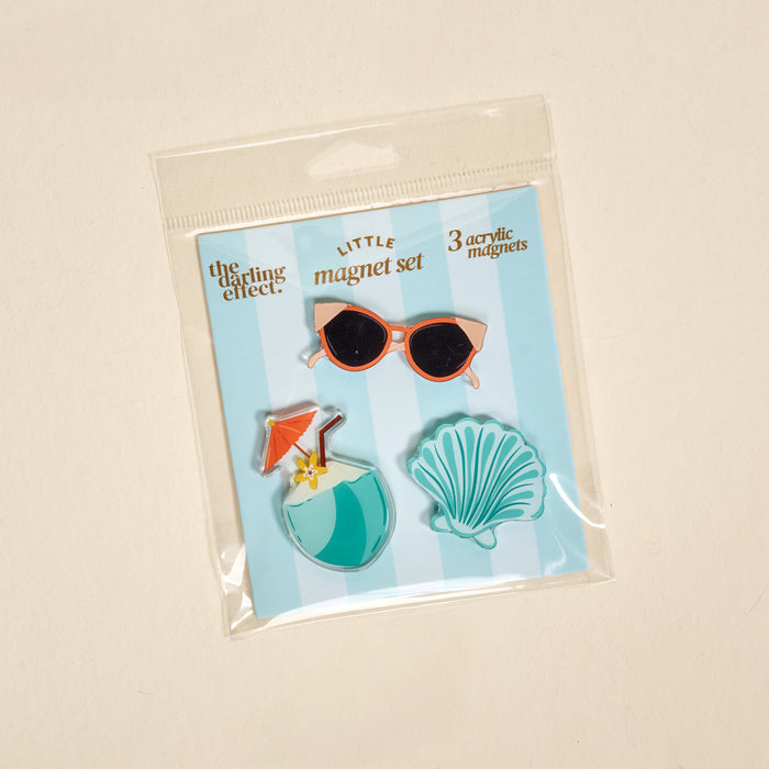 The Darling Effect Vacation Vibes Little Magnet Set features three acrylic fridge magnets—pink sunglasses, a tropical drink with an umbrella, and a teal seashell—packaged on a blue and white striped card in a clear box.