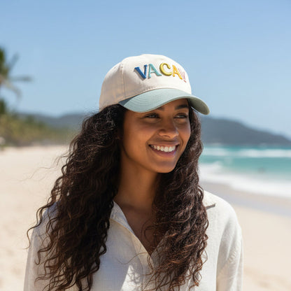A smiling woman with long curly hair wears The Darling Effect’s Vacay Classic Canvas Hat, featuring colorful "VACAY" letters, and a light shirt, standing on a sunny beach with ocean and hills in the background.