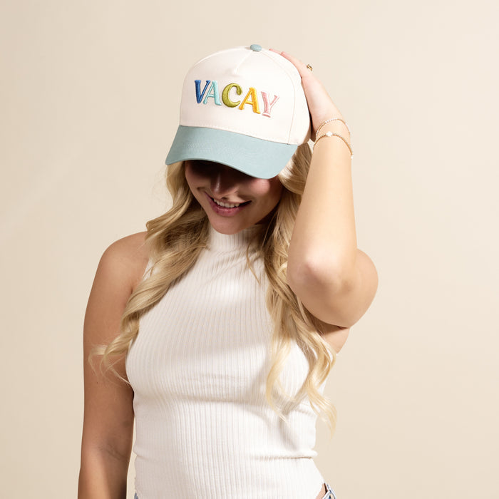 A woman with long blonde hair smiles while tilting The Darling Effect's Vacay Classic Canvas Hat—a white baseball cap with colorful "VACAY" lettering—pairing it effortlessly with a sleeveless white ribbed top against a beige background.