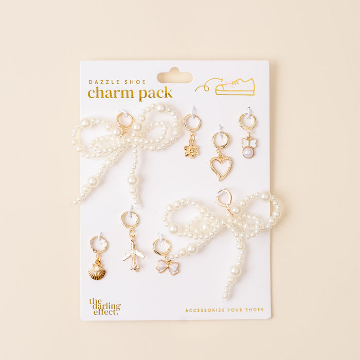 The Wanderlust Dazzle Shoe Charm Pack by the-darling-effect features a pearl bow and six gold charms—shell, airplane, star, heart, lock, and hexagon—displayed on a beige card for stylish footwear flair.