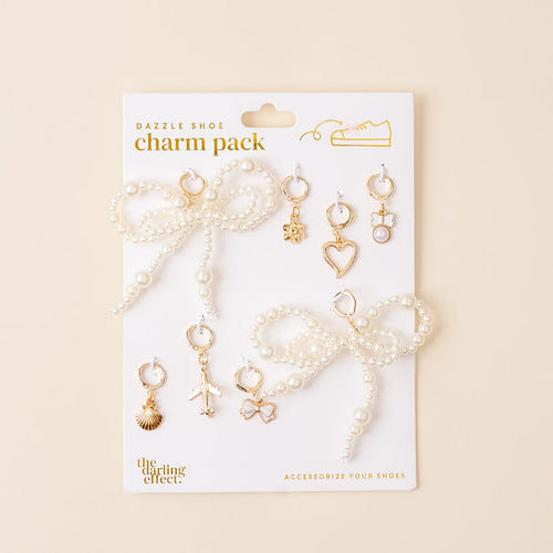 The Wanderlust Dazzle Shoe Charm Pack by the-darling-effect features a pearl bow and six gold charms—shell, airplane, star, heart, lock, and hexagon—displayed on a beige card for stylish footwear flair.