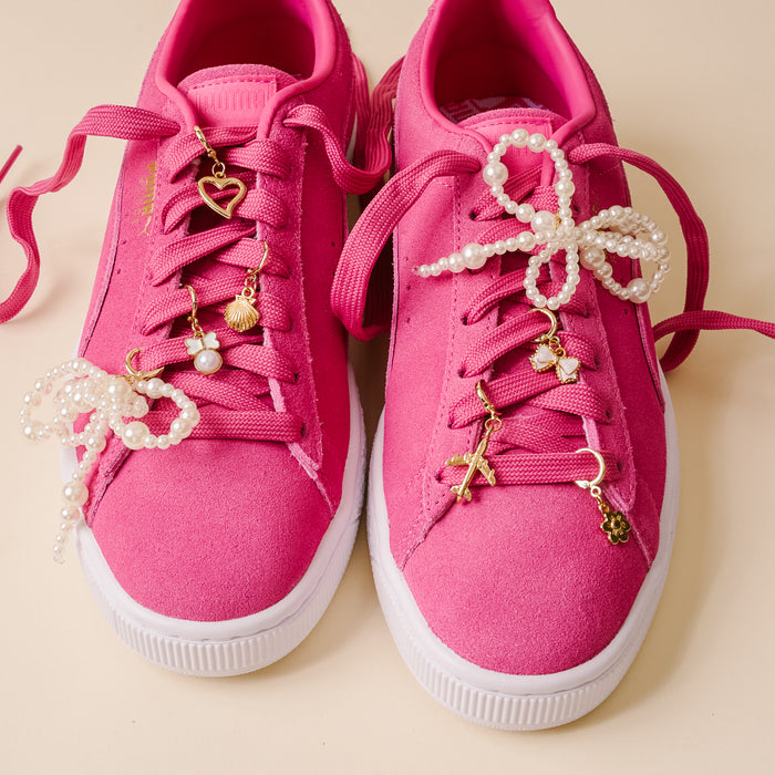 A pair of bright pink sneakers with white soles, pink laces, and the-darling-effect's Wanderlust Dazzle Shoe Charm Pack, featuring pearl beads and gold heart, cross, and flower charms.