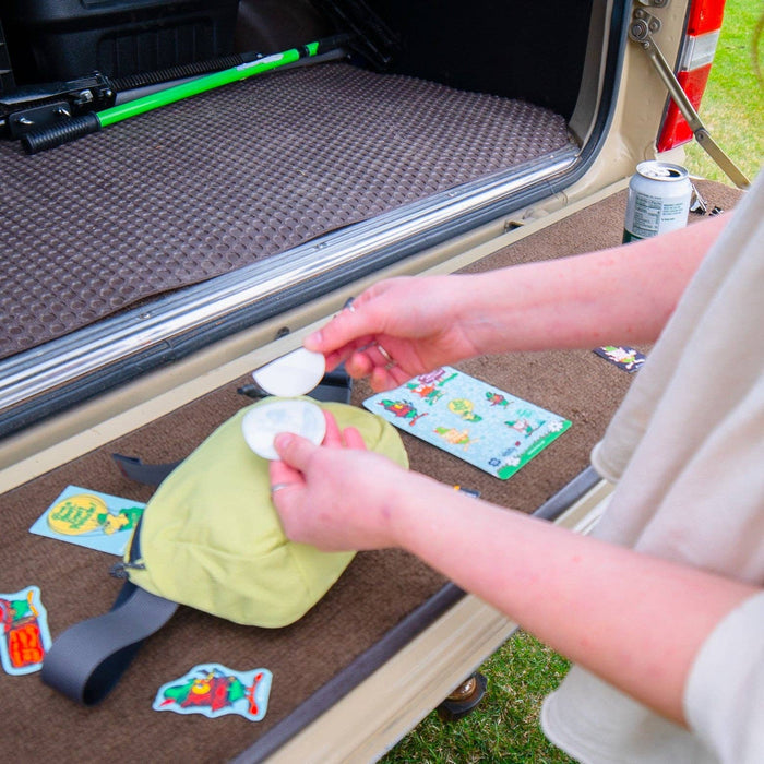 Hand holds Whistle Woodsy Owl NoSo Repair Patches by Noso near yellow bag on stickered car trunk; outdoor gear accessory.