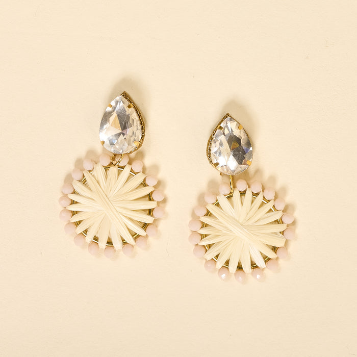 The Darling Effect's White Radiant Gem Earrings feature lightweight boho style with large teardrop clear gems atop circular woven details bordered in pale pink beads, shown against a cream background.