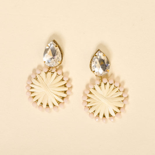 The Darling Effect's White Radiant Gem Earrings feature lightweight boho style with large teardrop clear gems atop circular woven details bordered in pale pink beads, shown against a cream background.
