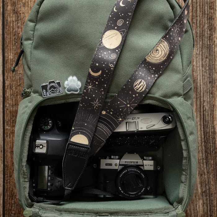 Open green backpack displays two cameras, Wildtree Sun & Moon Camera Strap, and three unique pins on its flap—perfect travel gear.