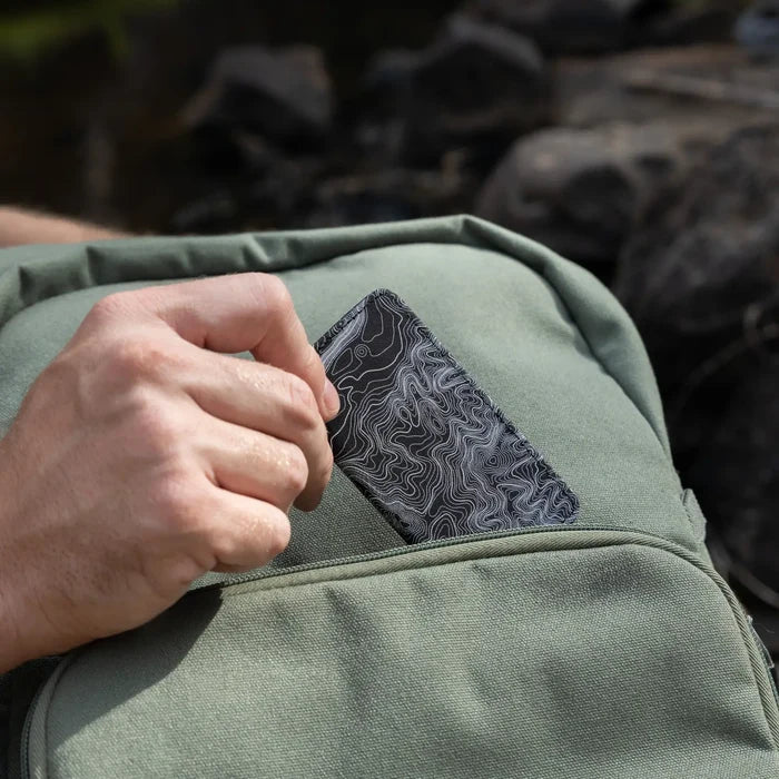Hand placing black Wildtree Topographic Map Switch card holder into green backpack front pocket outdoors, travel gear.