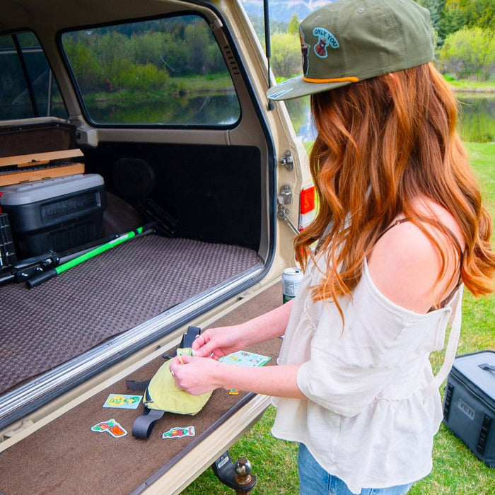 Applying a Wisdom Woodsy Owl x NoSo Repair Patch to a yellow camping bag by an SUV trunk during outdoor adventure.