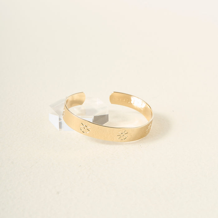 The Darling Effect’s You Are Outgoing Cuff Bracelet features a hammered gold finish with sunburst engravings, shown on a light surface beside a clear acrylic block—an elegant accessory and meaningful gift.