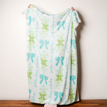 A person stands behind The Darling Effect's Zeta Tau Alpha Plush Sherpa Blanket, featuring light blue bows, green leaves, and cursive lettering. Only their hands and feet are visible against a wooden floor and white wall.